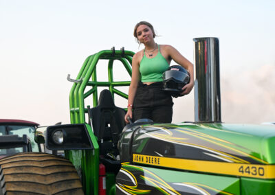 A young woman holds her helmet and stands on her tractor