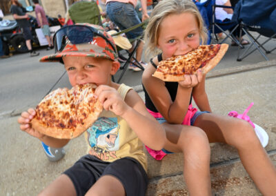 Brother and sister enjoying a large slice of pizza together at a downtown event, smiling and having fun.