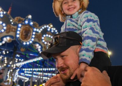 Young daughter on her father's shoulders with a carnival ride in the background at the Monroe County Fair.