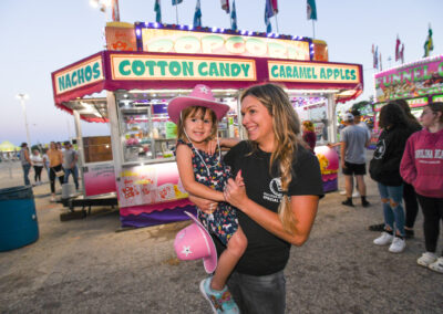 Young girl in a cowboy hat, smiling and held by her mother at a fair.