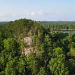 A picture of a bluff in summertime with a lake and forest behind it.