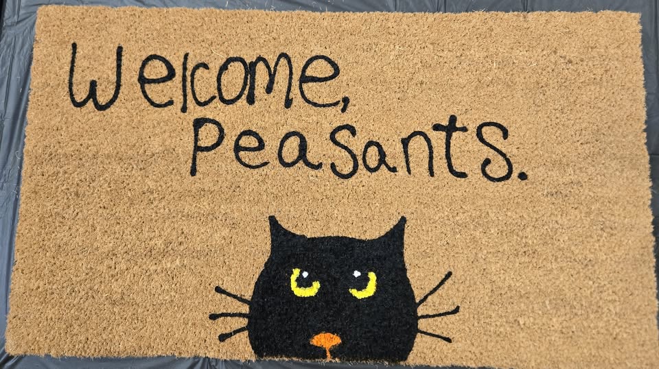 a doormat with the words "welcome peasants" above a black cat